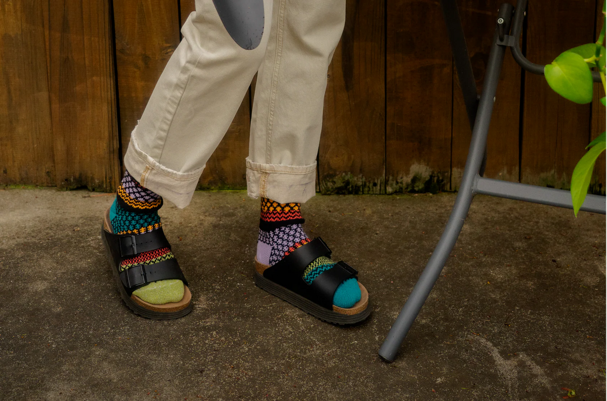 A person wearing black slide sandals with colorful Solmate patterned socks and a green toe cap, standing outdoors on a concrete surface in front of a wooden fence.