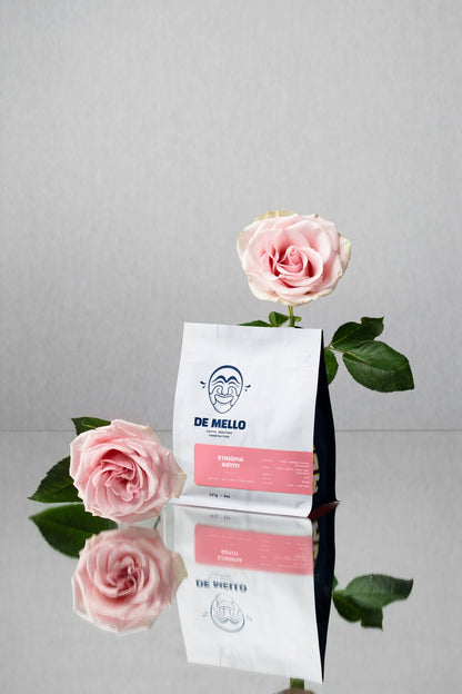 White coffee bag with pink label featuring 'De Mello' brand name, surrounded by pink roses on a reflective surface.