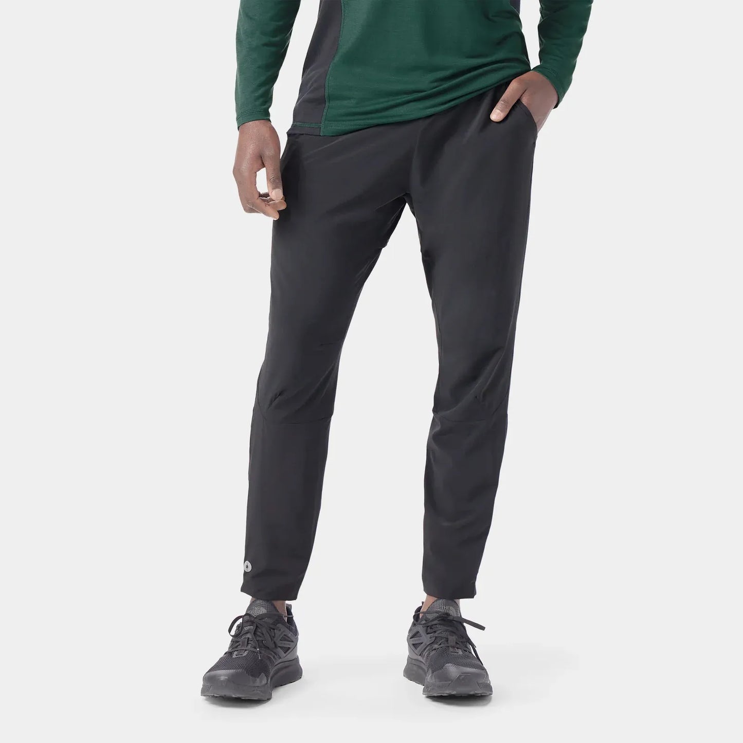 A person Smartwool Active Running pants with a green long-sleeve top and dark running shoes, shown from the waist down against a light gray studio background.