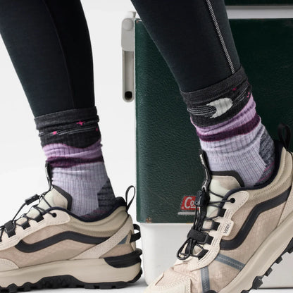 Close-up of a person's legs wearing Smartwool patterned crew socks and beige Vans hiking shoes with black accents, standing next to a dark green textured box.