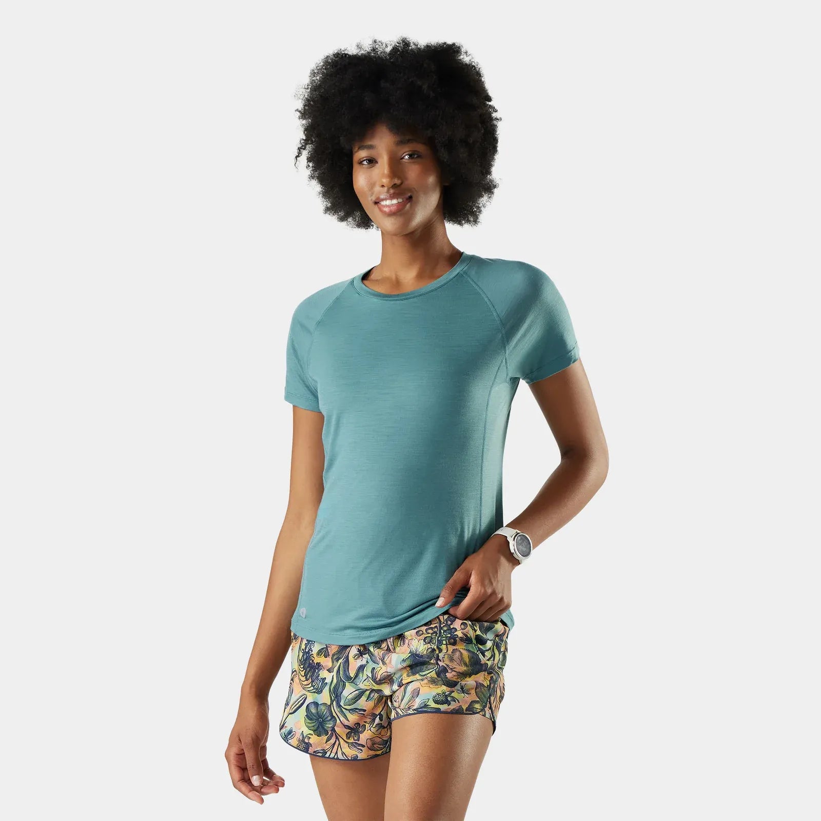 Woman wearing a teal Smartwool Active Ultralite shirt and floral shorts on a white background