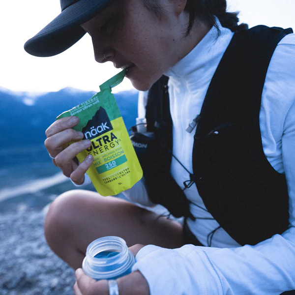 Person outdoors, tearing open a Naak Ultra Energy drink package with mountains in the background